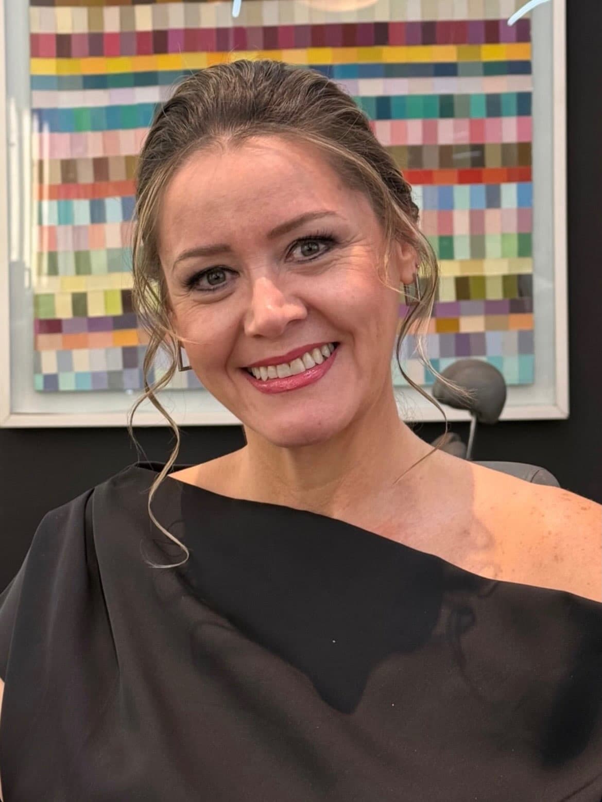 Cliente sorrindo com maquiagem marcante e coque delicado, diante de um quadro colorido