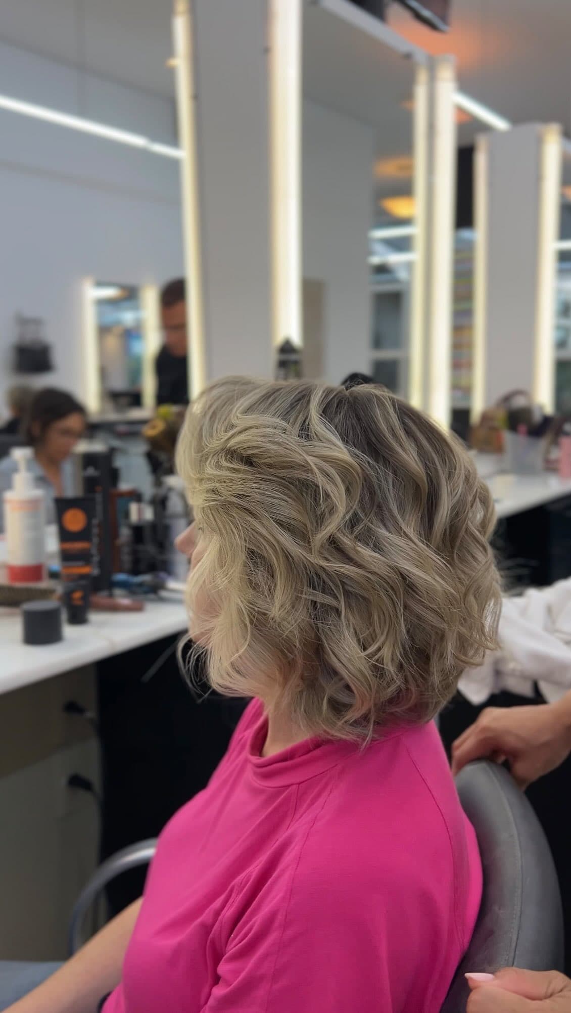 Cabelo curto loiro com ondas, cliente sentada diante dos espelhos com luz do salão