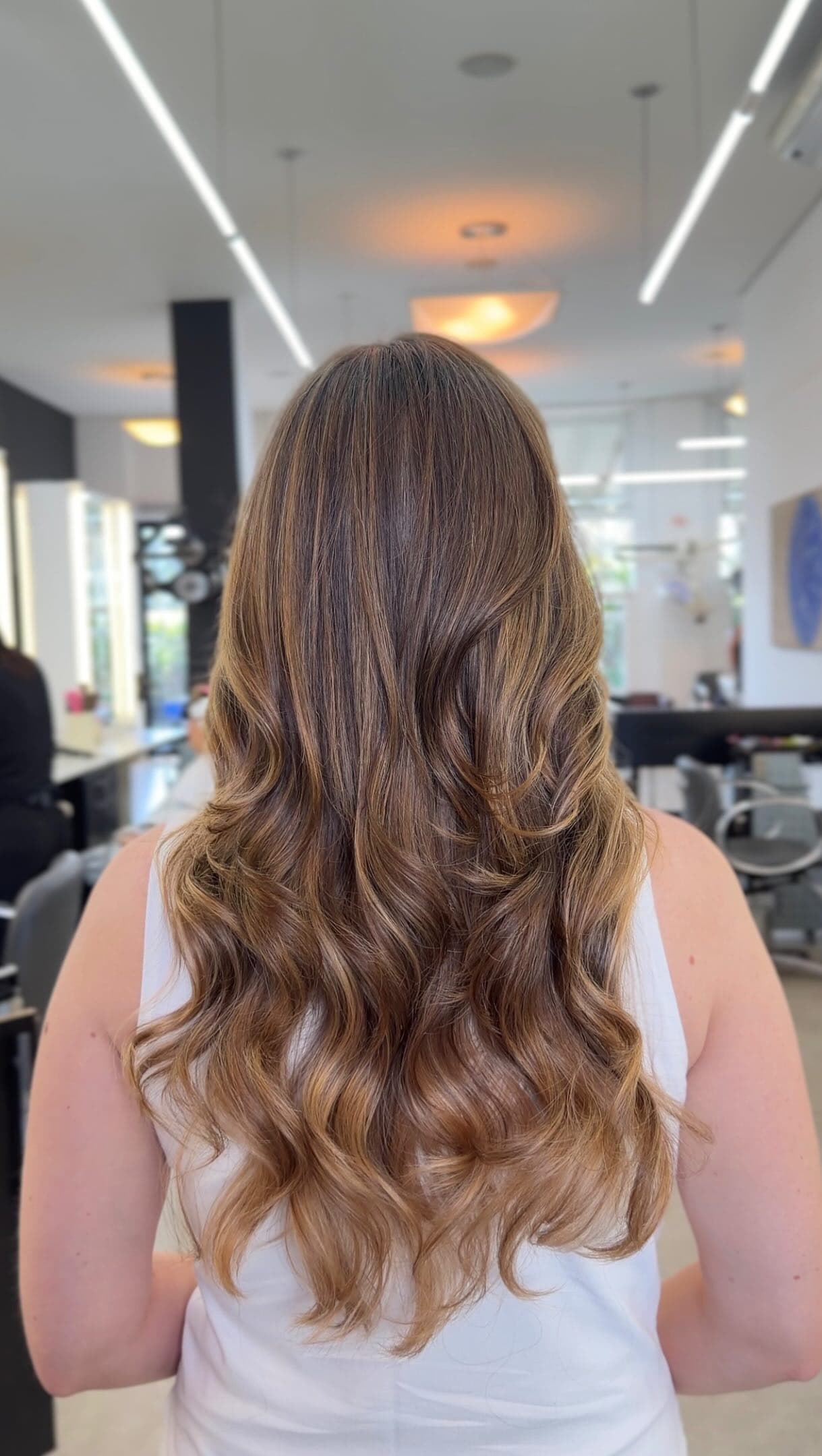 Cabelo longo castanho com mechas douradas e ondas, visto de costas no corredor do salão