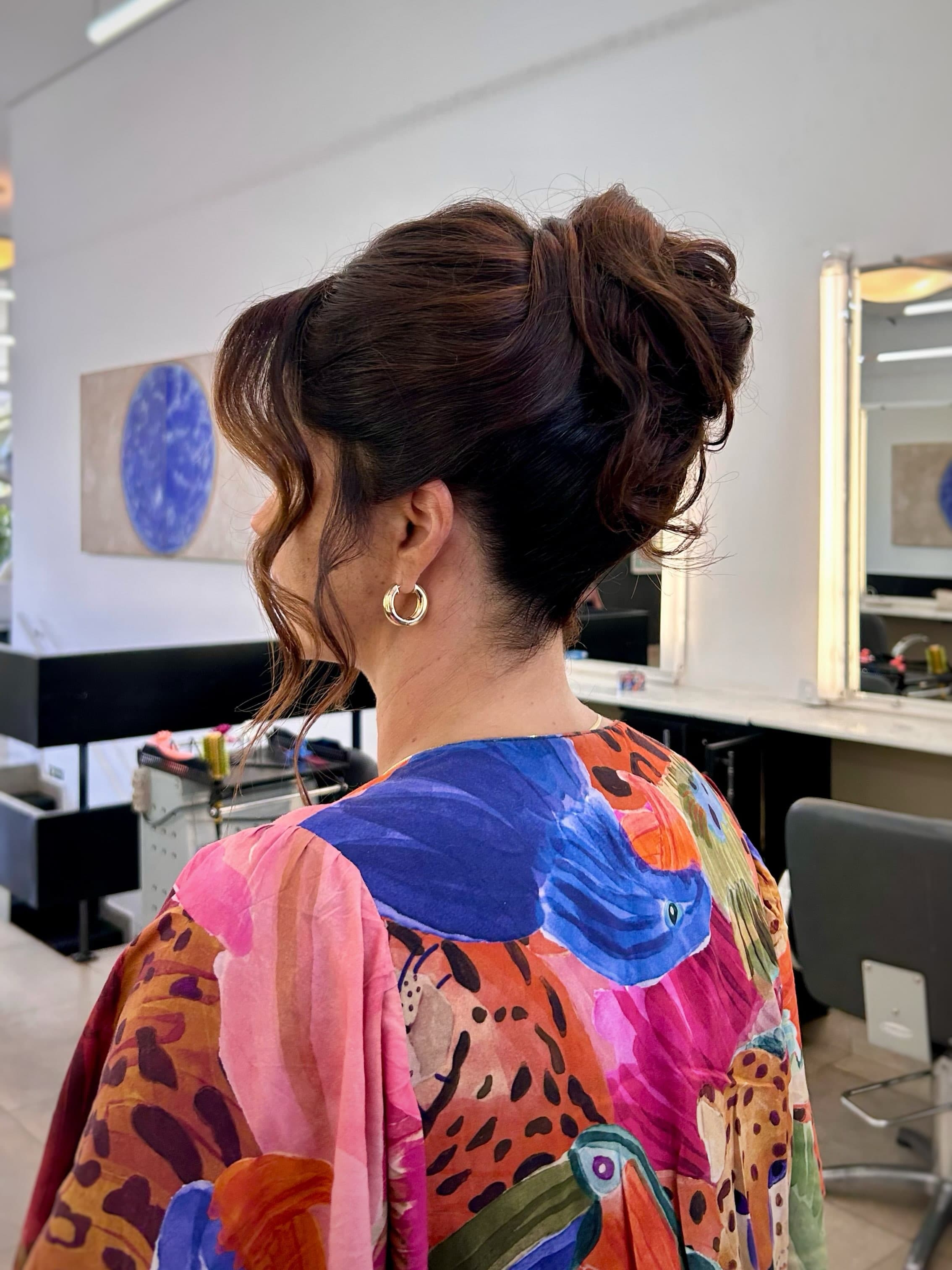 Coque alto com mechas soltas, de perfil, com o quadro azul redondo do salão ao fundo